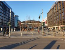 Straßenansicht mit der Mercedes-Benz Arena im Hintergrund, umgeben von modernen Gebäuden mit Fassaden aus Glas und Backstein. Im Vordergrund Fußgängerzone mit einigen Menschen, Ampeln und Sitzbereichen bei Cafés auf der rechten Seite.