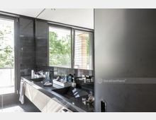 Modernes Badezimmer mit einer breiten, dunklen Waschtischfläche und zwei Waschbecken. Große Fenster lassen Tageslicht herein und bieten Blick auf grüne Baumkronen und eine Holzverkleidung außen.