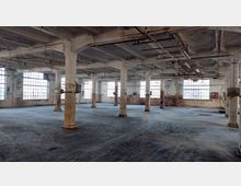 Große, leere Industriehalle mit sichtbaren Betonstützen und veralteter Bausubstanz. Die Wände sind teils abgeblättert, und große Fenster sorgen für Tageslicht; an der Decke verlaufen Rohre und alte Leitungen.