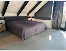 Schlafzimmer in einem Dachgeschoss mit schräger, weiß gestrichener Holzdecke und schwarzen Querbalken. Im Raum befinden sich ein großes Bett mit grauer Tagesdecke, ein kleiner Nachttisch mit Lampe, bodentiefe Fenster und dunkle Vorhänge.