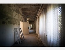 Ein verlassener, heruntergekommener Flur mit abgeblätterter Farbe an den Wänden und der Decke. Rechts befinden sich Fenster mit weißen, durchscheinenden Vorhängen, und auf dem Boden liegen Schutt und abgefallene Putzstücke.