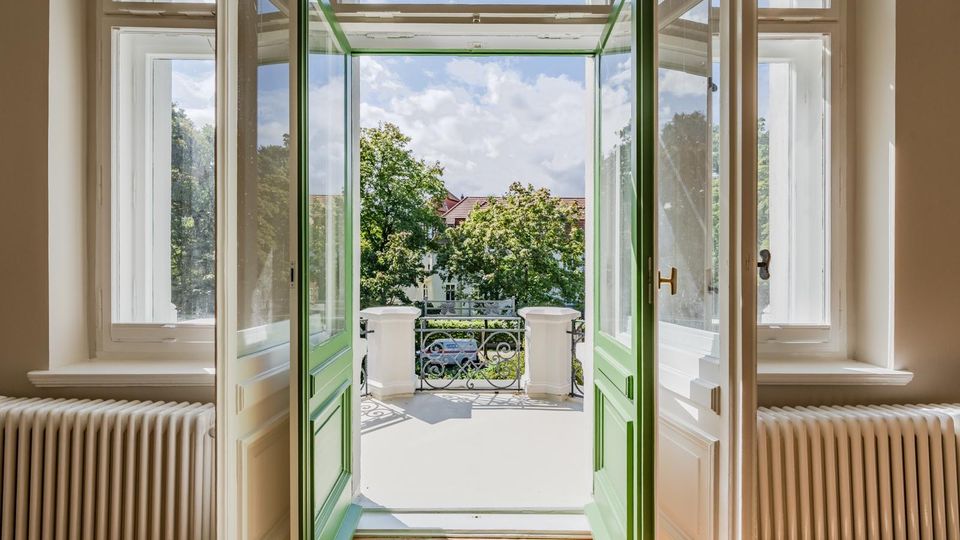 Blick aus einem hellen Raum durch doppelflügelige, grün gerahmte Fenstertüren auf einen kleinen Balkon mit schmiedeeisernem Geländer. Draußen sind Bäume, gegenüberliegende Wohnhäuser und ein blauer Himmel sichtbar.