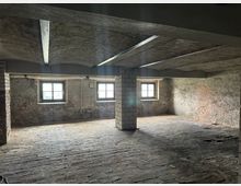 Ein dunkler Kellerraum mit rohem Mauerwerk und Ziegelboden. Drei kleine Fenster mit gitterartigen Rahmen lassen natürliches Licht herein, und massive rechteckige Säulen stützen die niedrige Decke.