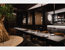 Innenansicht eines modernen Restaurants mit dunkler, eleganter Einrichtung. Im Vordergrund steht ein großer Baum in einem bepflanzten Kiesbereich, umgeben von langen, gedeckten Tischen und minimalistischen Holzstühlen; im Hintergrund sind beleuchtete goldfarbene Gitterelemente und eine Bartheke mit Glasregalen sichtbar.