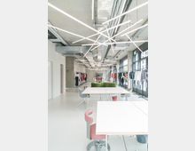 Heller, moderner Innenraum eines Showrooms mit offenen Decken, Sichtinstallationen und diagonal verlaufenden LED-Leuchten. Im Raum befinden sich weiße Tische, farbige Stühle und Kleiderständer mit Kleidung entlang der Wände.