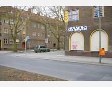 Straßenszene in einem Wohnviertel mit mehrstöckigen Putzbauten in brauner Farbe und gelben Fensterrahmen; im Vordergrund ein Gebäude mit geschlossenen Rollläden, beschriftet mit „Savan Elektro GmbH Berlin“, sowie Bäume und geparkte Autos. Verkehrszeichen und ein orangener Mülleimer stehen am Gehweg.