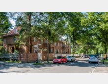 Straßenszene in einem Wohngebiet mit mehrstöckigen Backsteinhäusern, umgeben von hohen Bäumen und Grünflächen. Geparkte Autos säumen die Straße, die von einem Gehweg und einer niedrigen Backsteinmauer begrenzt wird.