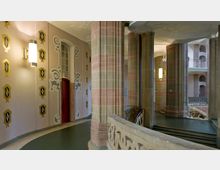 Innenansicht eines historischen Gebäudes mit kunstvollen Säulen aus sandfarbenem Stein und geschwungenen Treppengeländern. Die Wände sind mit geometrischen gold-schwarzen Ornamenten dekoriert, und eine rote Tür sowie Wandlampen prägen den Eingangsbereich.