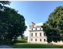 Ein weißes, historisches Gebäude mit klassizistischen Elementen, umgeben von Bäumen und einer Rasenfläche. Auf dem Turm des Gebäudes weht eine deutsche Flagge unter klarem, blauem Himmel.