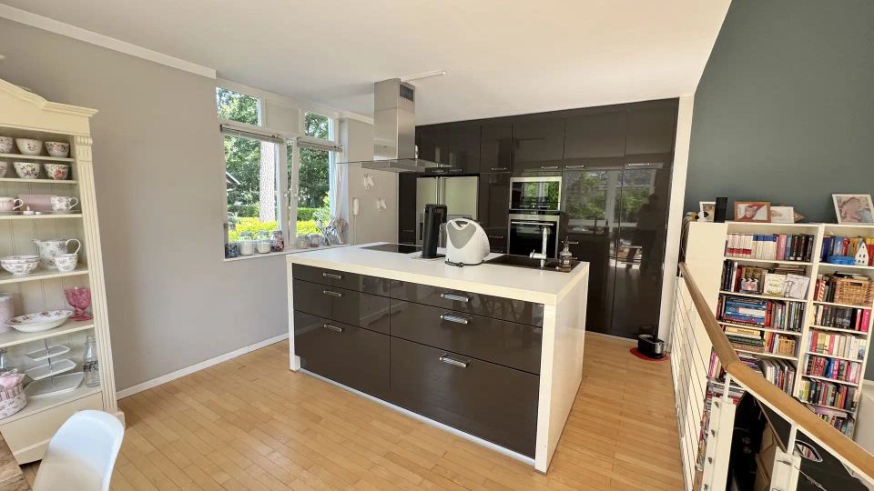 Moderne Wohnküche mit großer Kücheninsel, dunklen Hochglanzschränken und integrierten Backöfen, hellen Arbeitsflächen und Holzfußboden. Große Fenster mit Blick ins Grüne sowie angrenzende Regale und eine kleine Treppe auf der rechten Seite.