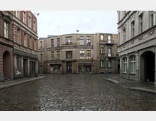 Pflastersteingasse mit historischen Gebäuden im Stil des frühen 20. Jahrhunderts, darunter eine Druckerei und Geschäfte mit verblassten Fassaden und Schaufenstern; mehrere Laternen säumen die Straße, die von feuchtem Regen reflektiert wird.