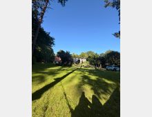 Große, sonnige Rasenfläche in einem baumbestandenen Garten mit langgezogenen Schatten, im Hintergrund ein niedriges Haus mit wellenförmigem Dach und üppigen Beeten. Rechts am Rand stehen geparkte Autos neben Büschen und Bäumen unter klarem blauem Himmel.