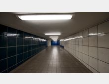 Ein unterirdischer Fußgängertunnel mit gefliesten Wänden, links in dunkelblau und rechts in weiß. Der Boden besteht aus grauen Fliesen, die Decke ist mit mehreren rechteckigen Leuchtstofflampen beleuchtet.