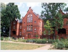 Backstein-Gebäude im neugotischen Stil mit Spitzbogenfenstern und dekorativen Zierelementen, umgeben von einem parkähnlichen Garten mit Wegen, Bänken und Bäumen.