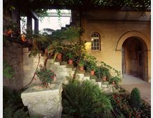 Steinarchitektur mit einem offenen Innenhof, der von Pflanzen und Blumentöpfen gesäumt ist. Eine Steintreppe mit Topfpflanzen führt nach oben, daneben befinden sich ein Rundbogen und ein Fenster in einer gelben Sandsteinwand.