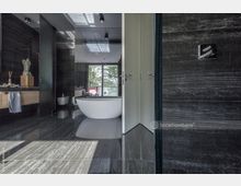 Modernes Badezimmer mit dunklen Marmorböden und -wänden, freistehender weißer Badewanne vor einem großen Fenster, das viel Tageslicht hereinlässt. Im Raum befinden sich außerdem ein wandmontiertes Waschbecken mit Holzunterschrank und ein Bidet, ergänzt durch minimalistische Beleuchtung.