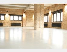 Industrie-Loft mit freiliegenden Backsteinwänden, großen schwarzen Fenstern und einem hellen, glatten Betonboden. An den Wänden hängen schwarze Heizkörper, und die Decke zeigt sichtbare Betonträger.