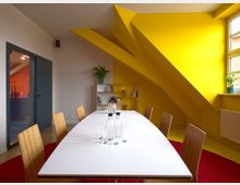 Ein moderner Besprechungsraum mit einem langen weißen Tisch, umgeben von Holzstühlen, auf rotem Teppichboden. Gelbe Wände mit schrägen Architekturelementen, ein Regal mit Pflanzen und Büchern, sowie ein Fenster mit natürlichem Licht prägen den Raum.