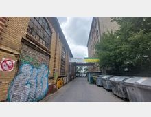 Enger Durchgang zwischen zwei Backsteingebäuden mit großen, bogenförmigen Fenstern; links markante Graffiti an der Wand, rechts eine Reihe von Müllcontainern und Bäume, oben ein Schild mit Firmenbeschriftungen über dem Weg.