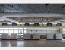 Abfertigungsschalterbereich eines Flughafens mit mehreren leeren Schaltern und nummerierten Schildern (B33 bis B38) über den Schaltern. Der Raum hat eine metallische Decke mit geometrischem Muster und eine helle Fensterfront im Hintergrund.