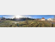 Weitläufiges, offenes Grundstück mit Grasbewuchs und einigen kahlen Bäumen, umgeben von Gebäuden, darunter ein gelbes Wohnhaus mit rotem Dach rechts und eine Reihe mehrstöckiger Wohnhäuser im Hintergrund. Der Bereich ist teilweise durch Bauzäune abgegrenzt, und die Sonne scheint tief am Himmel.