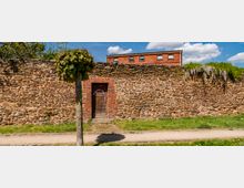 Eine historische Steinmauer mit einer kleinen Tür aus Holz in einem gemauerten Ziegelrahmen, davor ein schmaler Weg und eine Wiese mit einem Baum. Im Hintergrund ist ein rotes Backsteingebäude sowie blühender Blauregen zu sehen.