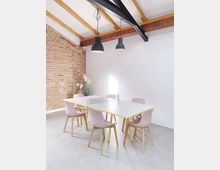 Ein moderner Besprechungsraum mit einer Tischtennistisch-Kombination, umgeben von rosa Stühlen mit Holzbeinen. Eine Ziegelwand, weiße Wände und eine Decke mit sichtbaren Holz- und Metallbalken prägen den Raum; zwei schwarze Hängelampen hängen von der Decke.