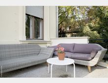 Terrasse mit zwei grauen Rattan-Sofas, eines mit lilafarbenen Kissen, und einem runden weißen Tisch, auf dem ein Blumentopf mit roten Blumen steht; im Hintergrund eine cremefarbene Hausfassade mit einem Fenster und umliegenden Bäumen.