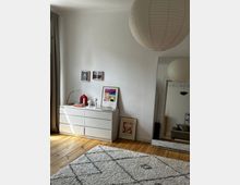 Helles Schlafzimmer mit Holzboden, weißer Kommode und großem Spiegel an der Wand. An der Kommode lehnen und hängen mehrere Bilder, ein Teppich mit geometrischem Muster bedeckt den Boden, und eine große, runde Papierlampe hängt von der Decke.