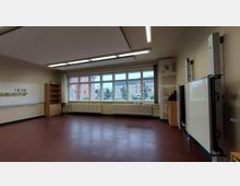 Ein leerer Klassenraum mit rotbraunem Boden, großen Fenstern an einer Wand, mehreren Heizkörpern darunter und Blick auf Gebäude außerhalb. An der rechten Seite befinden sich ein Whiteboard und ein Schrank, links an der Wand hängt eine leere Tafel.