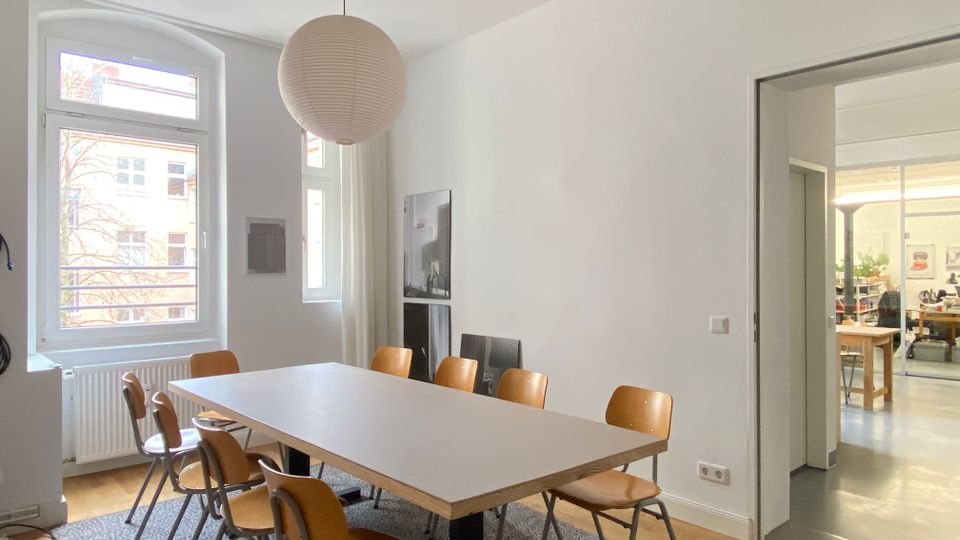 Heller Besprechungs- oder Essraum mit großem rechteckigen Tisch auf Teppich und mehreren Holzstühlen, beleuchtet von einer runden Pendelleuchte. Links große Fenster mit Blick nach außen und Heizkörper darunter; rechts eine breite Öffnung führt in einen angrenzenden Raum mit Werkstatt- oder Büroeinrichtung.