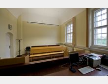 Ein kleiner Gerichtssaal mit hellgelben Wänden, Holzbänken und großen Fenstern mit Sprossen, die viel Tageslicht hereinlassen. Im Vordergrund befindet sich ein Schreibtisch mit einem Computer, einer Tastatur und einem Drucker.
