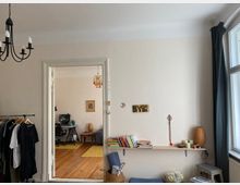 Wohnzimmeransicht mit einem Blick durch eine offene Tür in ein angrenzendes Arbeitszimmer. Im Vordergrund hängen Kleidungsstücke auf einer Stange, an der Wand ein Regal mit Büchern und Dekorationen, und rechts ein großes Fenster mit Vorhängen. Im Arbeitszimmer ein Schreibtisch mit Computer und weitere Möbel auf Holzboden.