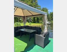 Überdachte Lounge-Ecke auf einer grünen Plattform am Wasser mit Rattan-Sofaecke, Sesseln und Glastisch unter einem Pavillon mit Vorhängen. Im Hintergrund sind ein ruhiger Teich oder Kanal, Uferbewuchs und Bäume zu sehen.