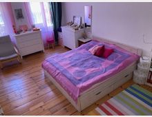 Ein Schlafzimmer mit einem Holzboden, einem weiß gestrichenen Bett mit lilafarbener Bettwäsche, mehreren dekorativen Kissen, einem Nachttisch, und weißen Kommoden. Der Raum ist hell beleuchtet durch Tageslicht, das durch Fenster mit rosa Vorhängen einfällt.