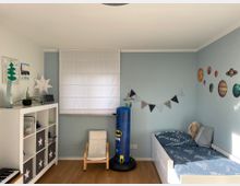 Kinderzimmer mit hellblauen Wänden und einem Holzboden. Links steht ein Regal mit Fächern und dekorativen Aufbewahrungsboxen, daneben ein kleiner weißer Sessel. Rechts ein Kinderbett mit blauer Bettwäsche, an der Wand darüber sind Wandsticker von Planeten angebracht. Ein weißer Rollo bedeckt das Fenster in der Mitte der Wand.