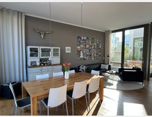 Wohnzimmer mit Essbereich, ausgestattet mit einem Holztisch und weißen Stühlen, einem grauen Sofa, einem schwarzen Ledersessel und einem weißen Teppich. Im Hintergrund befinden sich ein vintage Küchenschrank, eine Fotowand und große Fenster, die auf eine Terrasse mit Pflanzen blicken.