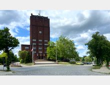 Backsteingebäude mit Turm und Uhr an der Fassade, umgeben von Bäumen und Grünflächen; davor eine breite Treppe und ein gepflasterter Platz mit parkenden Autos.