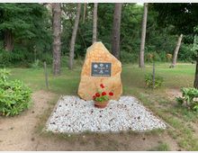 Gedenkstein in einem Park, umgeben von hohen Bäumen und Grünflächen. Der Stein steht auf einer mit weißen Kieselsteinen bepflanzten Fläche mit einem Blumentopf voller roter Blumen davor.