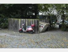 Ein roter Motorroller steht vor einer hölzernen Barriere, neben der eine weiße Abfallmulde lehnt. Im Hintergrund sind Bäume, Pflastersteine und parkende Autos zu sehen.