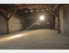Dunkler Dachboden mit Holzbalken und Holzboden, durch dessen kleines Fenster ein Lichtstrahl in den Raum fällt. Die Dachkonstruktion und die Wände wirken alt und unbehandelt.