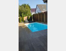 Privater Außenbereich mit rechteckigem Pool, umgeben von einer Holzterrasse und einem Holzzaun. Im Hintergrund befinden sich grüne Bäume, Büsche und zwei Liegestühle neben dem Pool.