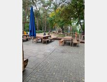 Außenbereich mit gepflasterter Terrasse und Holzbänken, daneben ein geschlossener blauer Sonnenschirm. Im Hintergrund sind ein Wald mit hohen Bäumen, ein Klettergerüst und spielende Kinder zu sehen.