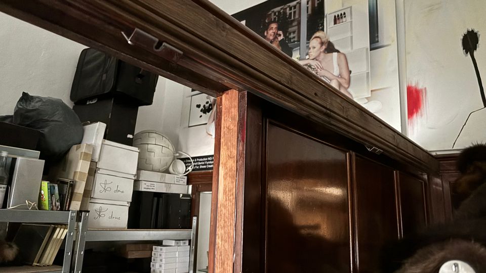 Innenraum eines Lager- oder Atelierraums mit hohen, dunkel vertäfelten Holzelementen und einem Metallregal voller Kisten, Taschen und Materialien. An der weißen Wand hängen große Fotodrucke und ein abstraktes Bild mit rotem Farbfleck und dunkler Blütenform.