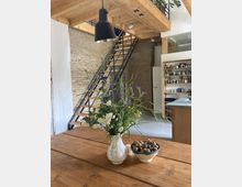 Holzgetäfelter Innenraum eines modernen Lofts mit sichtbaren Backsteinwänden. Im Vordergrund steht ein Holztisch mit einem Blumenstrauß in einer weißen Vase und einer Schale mit Nüssen; im Hintergrund eine Metall-Holz-Treppe mit Zwischengeschoss und Regalen.