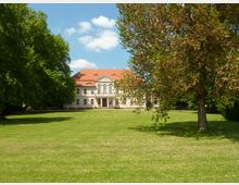 Herrenhaus in klassizistischem Stil mit hellen Fassaden und rotem Walmdach, im Hintergrund von Bäumen umgeben. Davor eine weitläufige, gepflegte Rasenfläche mit vereinzelten großen Bäumen.