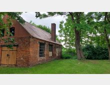 Backsteingebäude mit spitzem Ziegeldach und einem Schornstein, umgeben von Bäumen und Grünfläche; die Fassade zeigt eine Holz-Doppeltür und große Fenster. Im Hintergrund dichter Bewuchs und wolkiger Himmel.