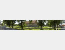 Panorama einer ländlichen Straße mit einer Reihe großer Bäume, die den Gehweg säumen. Im Hintergrund steht ein großes Gebäude aus Naturstein mit einem roten Satteldach, umgeben von einer umzäunten Wiese.