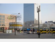 Aufnahme des Alexanderplatzes in Berlin mit der Weltzeituhr im Vordergrund, einem zylindrischen Monument mit internationalen Städtenamen und Uhrzeiten. Im Hintergrund sind ein modernes Hochhaus mit Glasfassade, Straßenbahnen, Geschäfte und Werbetafeln sichtbar.