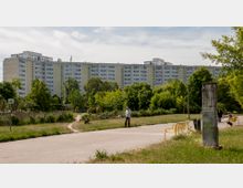 Ein Park mit Gehwegen, Sitzbänken und Grünflächen im Vordergrund, umgeben von Bäumen und Sträuchern. Im Hintergrund steht ein großes Mehrfamilienhaus in Plattenbauweise.